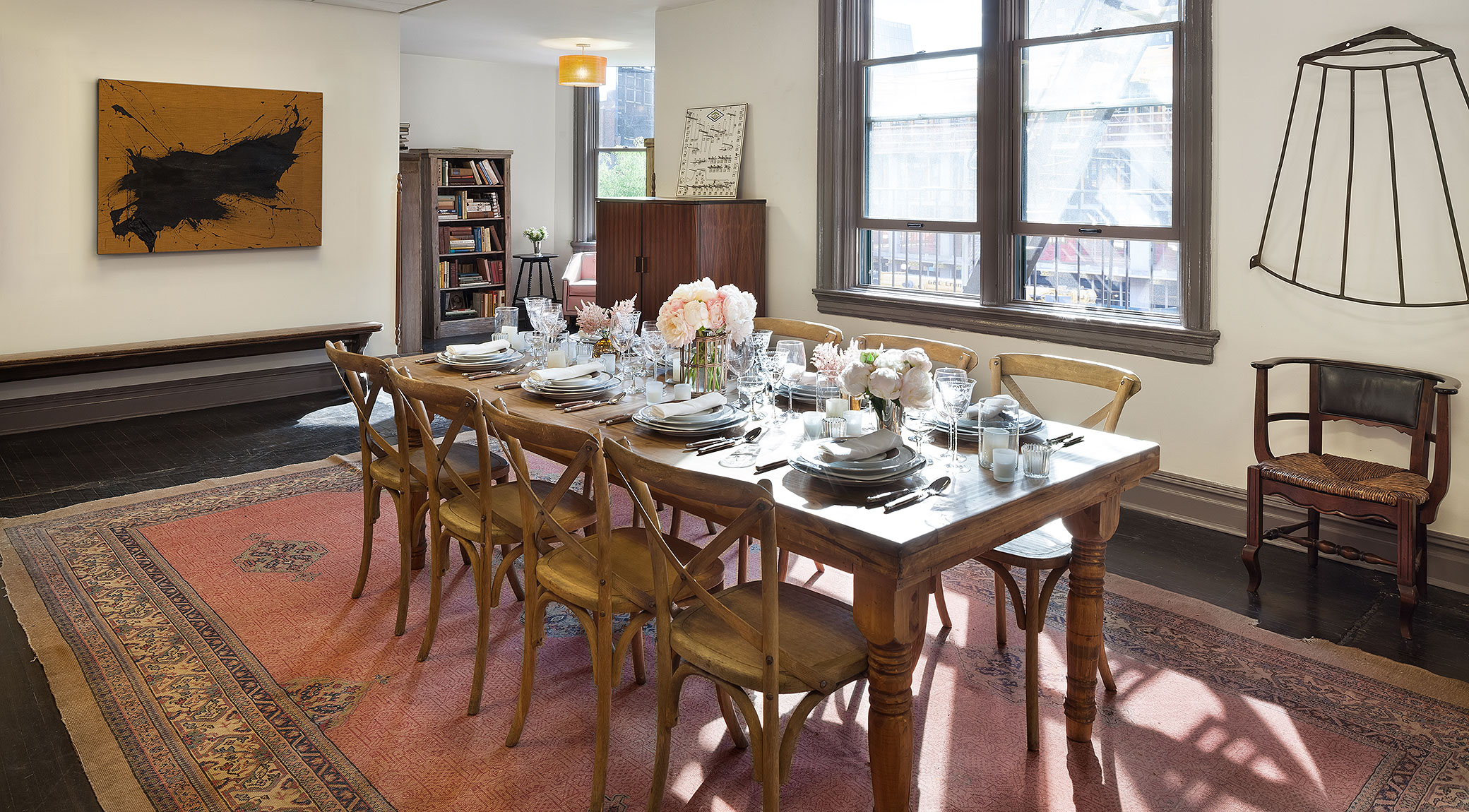 A dining room with a long wooden table set for a meal, surrounded by chairs, with a large rug underneath and artwork on the walls.