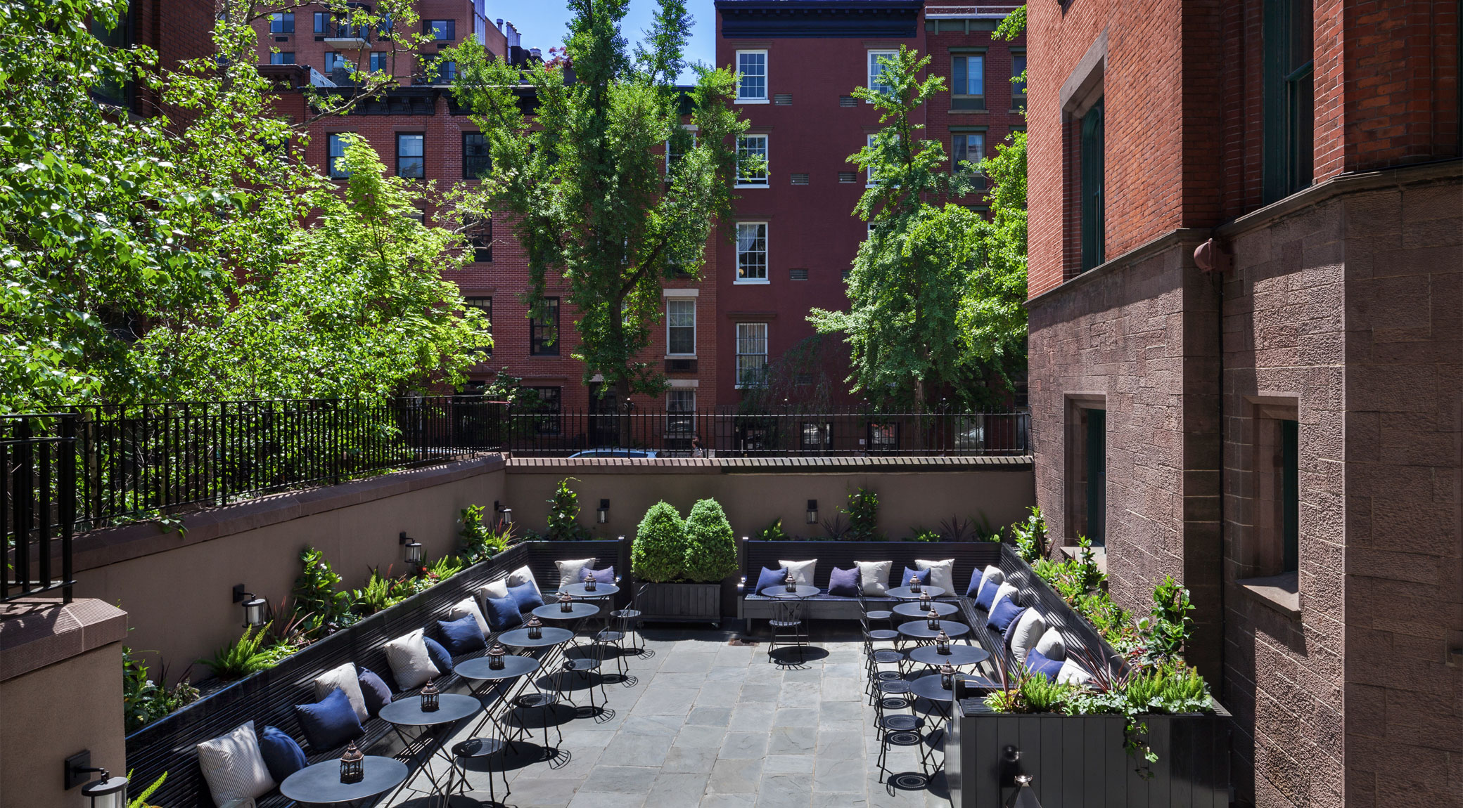A small outdoor seating area with tables, chairs, and pillows in a courtyard surrounded by buildings and trees.